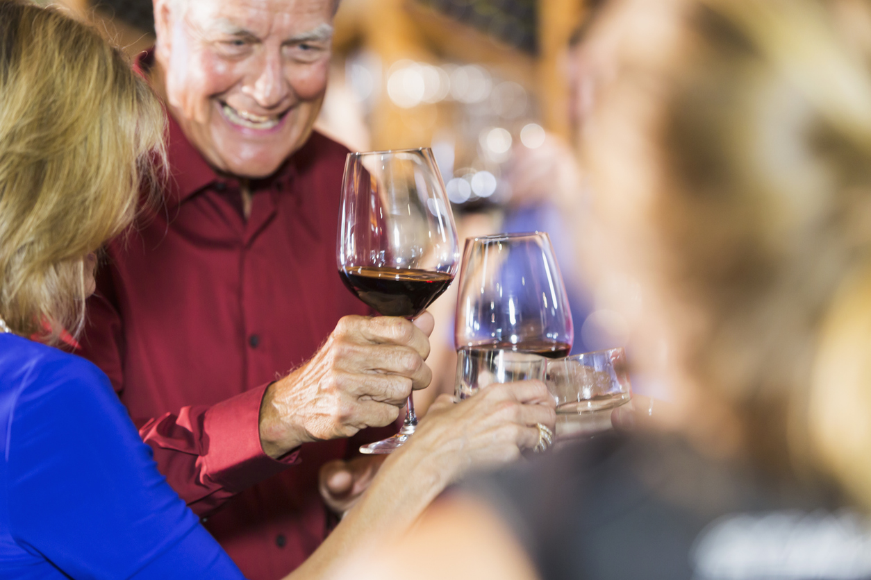 Close up, cropped view of a group of people at a party, drinking and toasting. The main focus is on the hand and wine glass of a senior man whose face is out of focus.