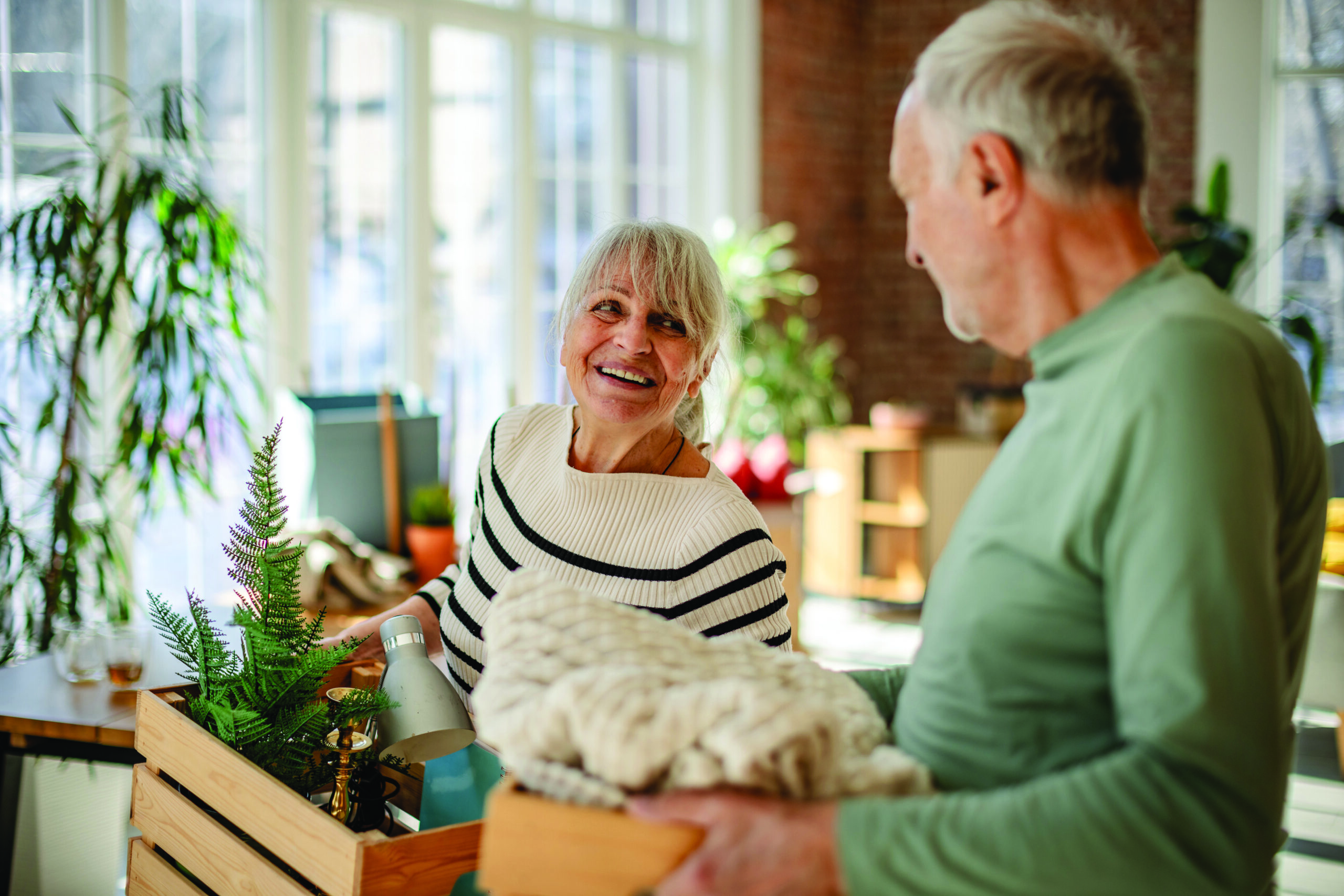 A senior couple is happily unpacking boxes together in their new living room, filled with plants and home decor. They share smiles as they arrange their belongings, enjoying their fresh start.